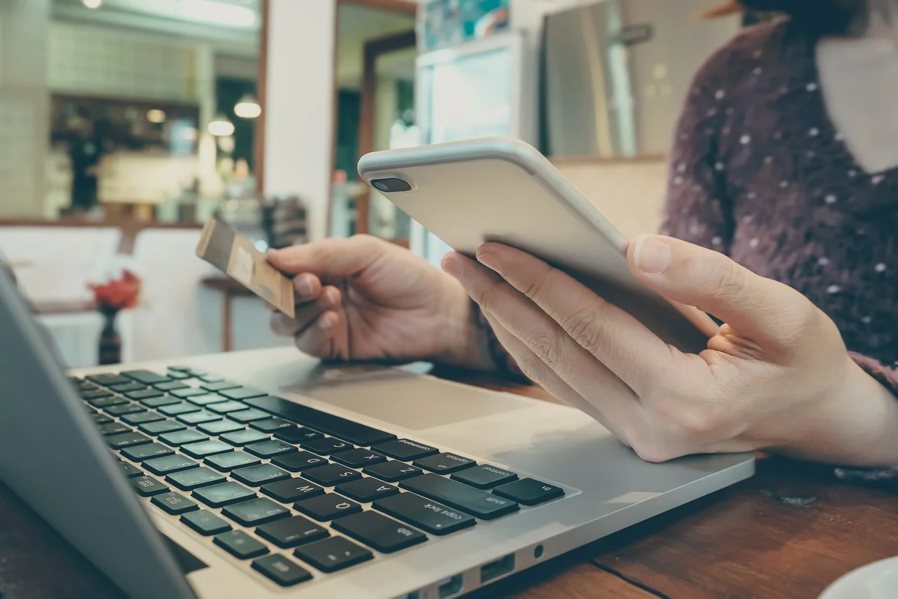 A person holding a phone and payment card beside a laptop